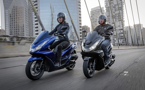 Duas motos em destaque na cidade, com pilotos usando capacetes e roupas de proteção, em uma estrada urbana moderna.