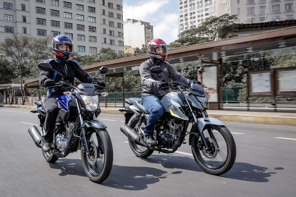 Duas motos em um passeio urbano, com pilotos usando capacetes coloridos, passando por um cenário de prédios.
