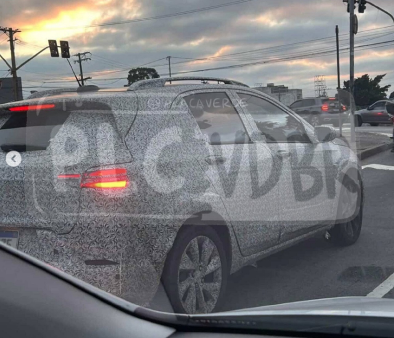 Protótipo de carro com camuflagem em rua movimentada, atrás de outro veículo, sob um céu nublado.